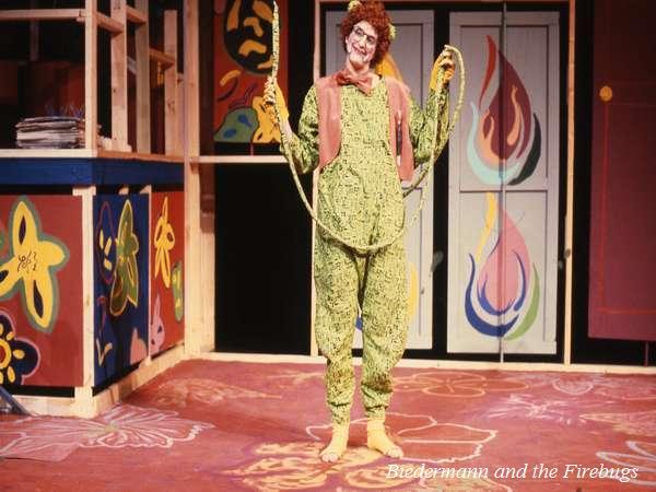 A performer in a green, patterned costume stands on stage holding a rope. The backdrop features colorful, abstract murals with flames and swirls. The scene is from "Biedermann and the Firebugs.