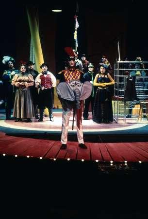 A person dressed in a colorful circus-like costume with an elephant face on their chest stands center stage. Behind, a group of people in theatrical costumes are gathered near a metal cage prop, set against a brightly lit backdrop.