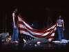 People holding a large American flag on a dimly lit stage. Some individuals are standing while others are lying on the ground, with the flag partially draped over them. The scene is dramatic and theatrical.