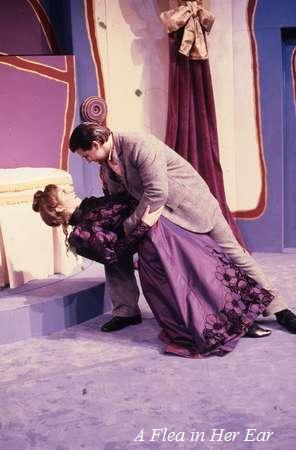 A man in a suit dramatically holds a woman in a purple dress who appears to faint or swoon in a theatrical setting. The stage has a colorful backdrop with abstract designs, and the text "A Flea in Her Ear" is visible at the bottom.