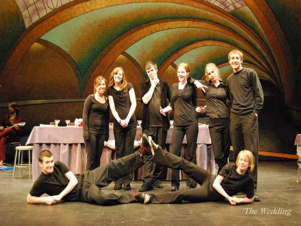 A group of eight people dressed in black performing on stage. Seven stand side by side, with two lying on the floor in front. The backdrop features colorful, curved ceiling designs. A table with plates and cups is visible behind them.