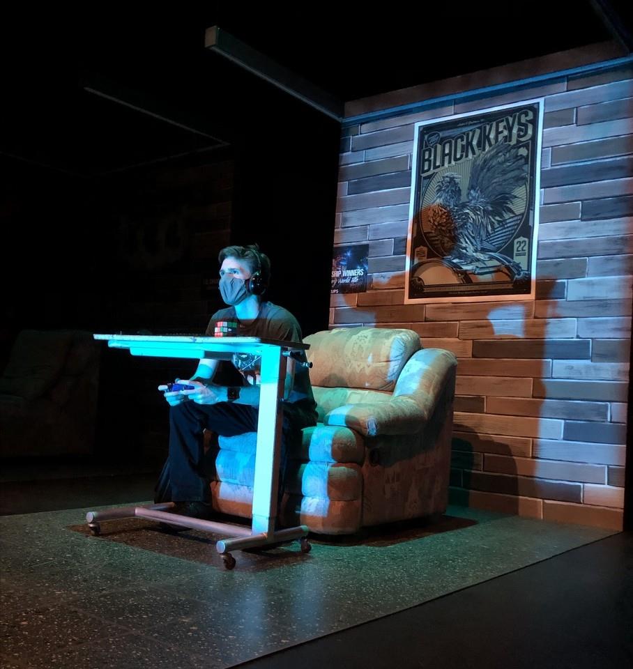 A person sits on a sofa, wearing headphones and holding a game controller. They face a TV screen, with a wooden wall behind featuring a Black Keys poster. The room is dimly lit, creating a focused atmosphere.
