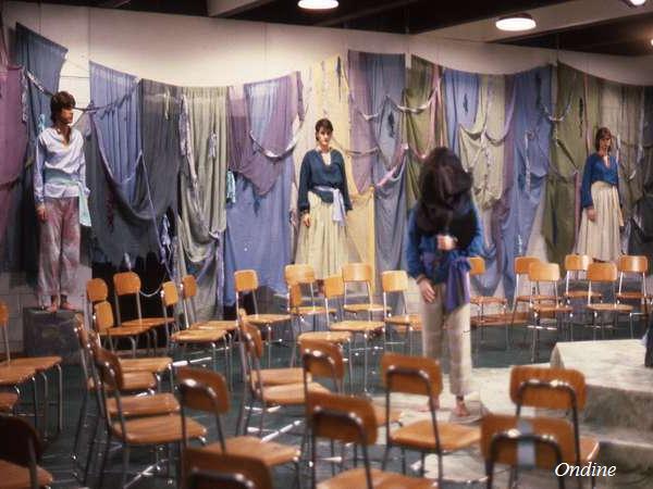 A theater stage with draped colorful fabrics in the background. Four performers in casual blue and white attire stand or move on stage. Empty chairs are arranged facing the stage. "Ondine" is written in the bottom right corner.