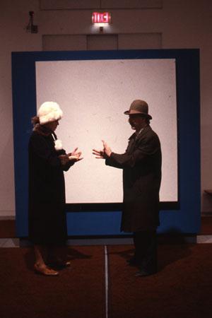 Two people in dark coats and hats stand facing each other in front of a large white square with a blue border on the wall. The room has a carpeted floor and an exit sign above the square.