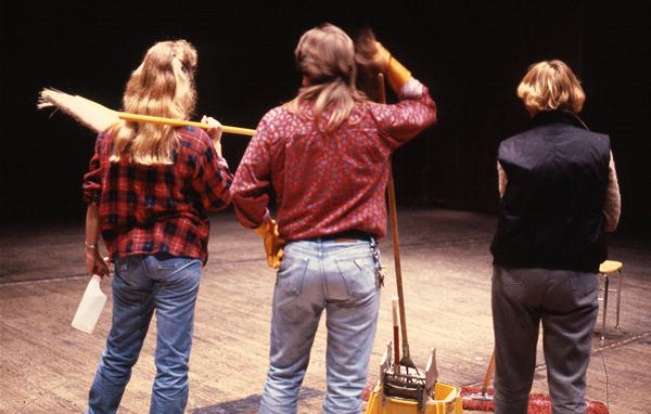 Three people stand with their backs to the camera on a stage. Two hold brooms and wear plaid shirts, jeans, and have long hair. The person on the right has short hair, a dark vest, and jeans. The stage is dimly lit.