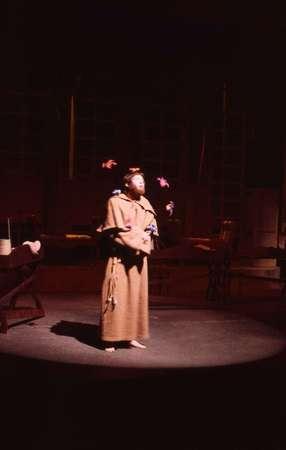 A person dressed in a long, brown robe stands on a stage under a spotlight, juggling three red balls. The background is dimly lit with wooden furniture partially visible.