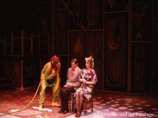 Three actors on stage in a theatrical scene. One, in a green costume, leans toward a seated man in a suit. A woman in a floral dress and hat sits beside them. The backdrop has dark panels with abstract designs. Text reads "Biedermann and the Firebugs.