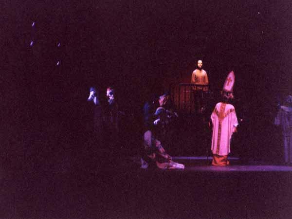 A dimly lit stage scene depicting several figures in medieval attire. One figure is standing in an elevated position, while others gather around below, focused on the central figure. The atmosphere is dramatic and theatrical.