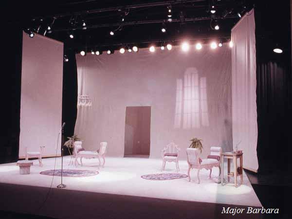 A theater stage set with minimal furniture including several chairs and a small table. The backdrop is plain with soft lighting creating a window-like shadow. The words "Major Barbara" are in the lower right corner.