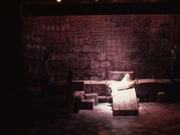 Dimly lit stage with a person dressed in white lying on a wooden platform in the center. Stone wall backdrop and scattered stage props add a mysterious atmosphere. Two shadowy figures are partially visible on either side.