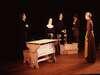 Five people dressed in period costumes stand around a wooden table on a stage with a dark background. They appear to be engaged in a serious discussion.