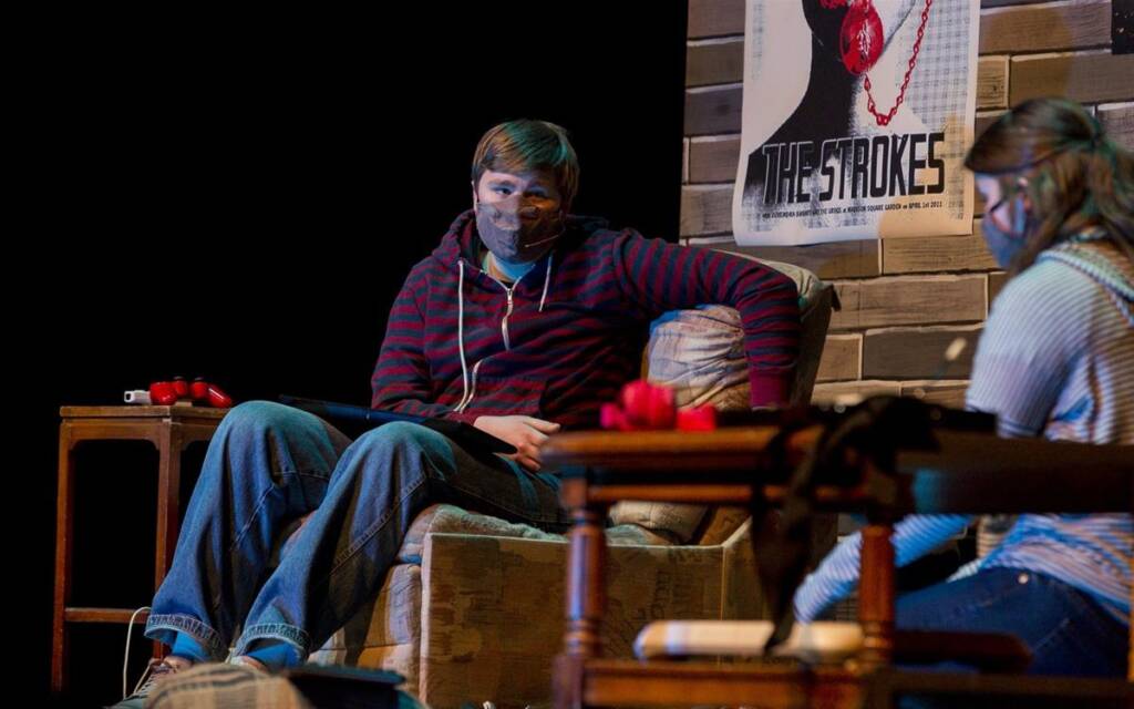 Two people sitting on stage in a play setup. One is on an armchair wearing a striped hoodie, and the other sits beside a table with red objects. A poster of "The Strokes" is on the wall behind them. Both are wearing face masks.