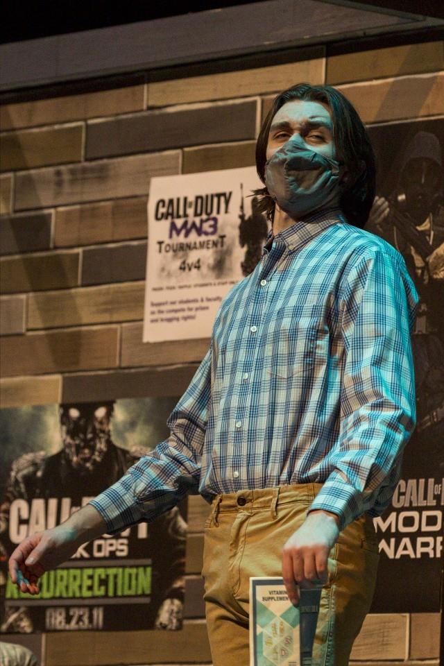 A person wearing a checkered shirt and a face mask stands in front of posters for Call of Duty and a tournament. They are holding a booklet and appear to be speaking or presenting. The setting suggests a gaming event or convention.