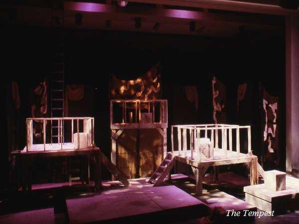 A dimly lit theater stage set for a production of "The Tempest." The set features wooden platforms and ladders, with purple and brown lighting casting dramatic shadows. The atmosphere is mystical and intense.