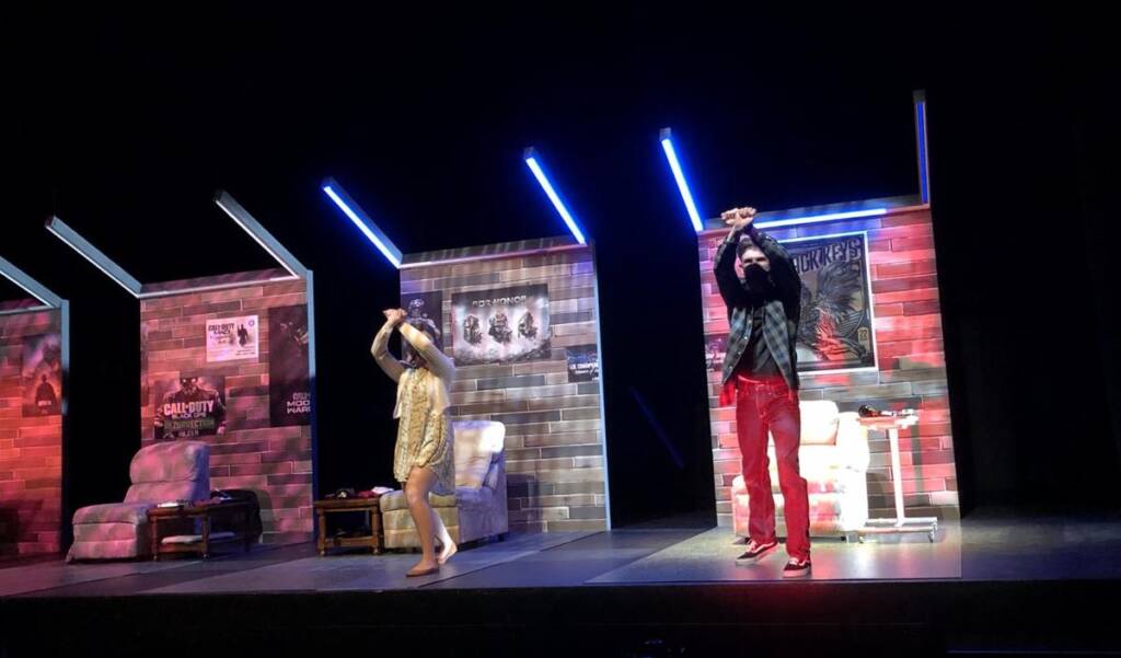 Two people on a stage are performing with lively expressions, raising their arms. The backdrop features posters and a living room setup with a couch and table. Bright lights illuminate the scene, creating a dynamic atmosphere.