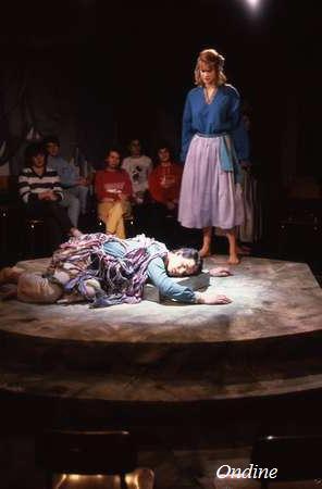 A seated audience watches a theatrical performance on a small stage. A person wearing a blue shirt and light purple skirt stands over another person lying on the ground, draped in fabric. "Ondine" is written in the bottom right corner.