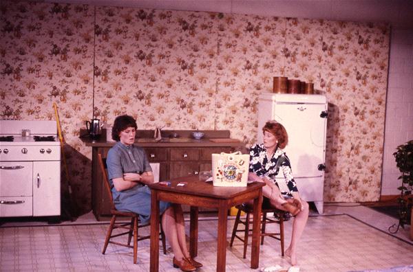 Two women sit at a small table in a vintage kitchen with floral wallpaper. One woman is wearing a blue dress, and the other is in a floral dress. A white refrigerator and stove are in the background. They appear to be engaged in conversation.