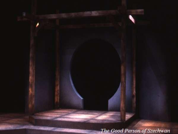 Dimly lit stage set for a play, "The Good Person of Szechwan," featuring a wooden frame with a circular doorway. The scene has muted colors and a minimalist design.