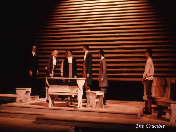 Five actors in historical costumes stand on a dimly lit wooden stage set designed to resemble a courtroom scene. A table and bench are centrally placed, with "The Crucible" written in the bottom right corner.