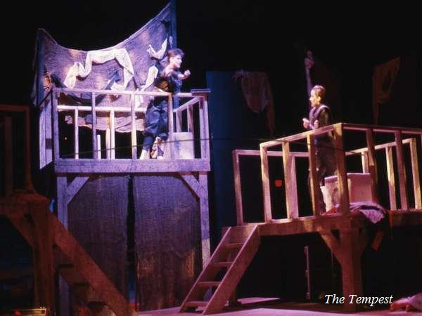 A stage scene from a play with two actors on elevated platforms. One actor gestures energetically while the other stands opposite them. The set includes wooden structures and fabric drapes. The words "The Tempest" are in the bottom right corner.