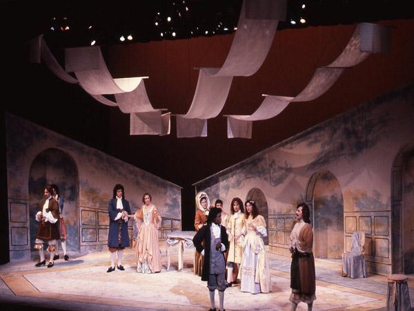 A theatrical performance on stage features actors dressed in period costumes. The set includes arched doorways and abstract backdrops. White fabric hangs from above, and the lighting casts warm tones across the scene.