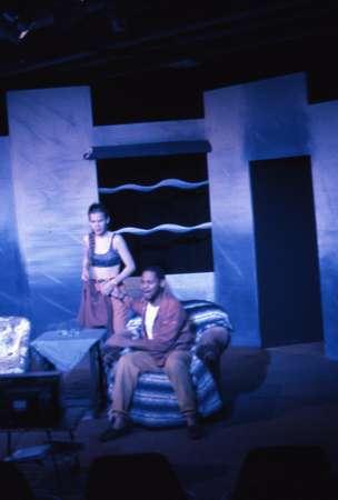 Two actors are on a stage set, designed to look like a living room with stylish, modern furniture. One actor sits on a chair, and the other stands nearby. The scene is lit in a blue hue, creating an atmospheric mood.