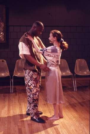 Two people stand closely facing each other on a wooden stage, surrounded by empty chairs. The person on the left wears a patterned outfit, and the person on the right wears a light-colored dress. They appear to be engaged in an intimate dance or theatrical scene.