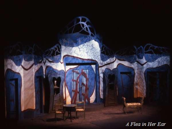 A stage set with a whimsical blue and white design featuring intricate patterns and abstract shapes. There's a small round table with chairs and a chaise lounge. The words "A Flea in Her Ear" are displayed in the corner.