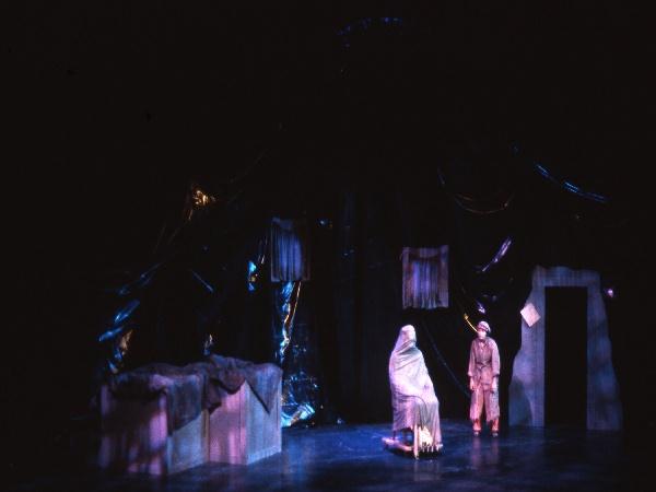 A dimly lit stage with two actors: one seated and cloaked, the other standing nearby. The background features a dark, abstract set design with draped fabrics and muted blue lighting.