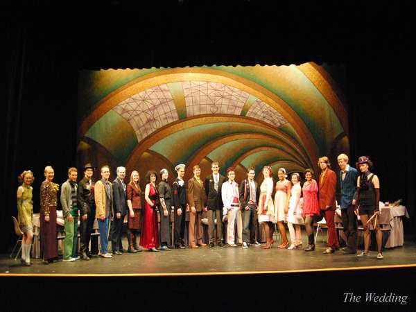 A group of people in colorful, vintage-style clothing stands on a stage with an ornate, arched backdrop. They are lined up side by side, posing for a photograph. The words "The Wedding" are visible in the bottom right corner.