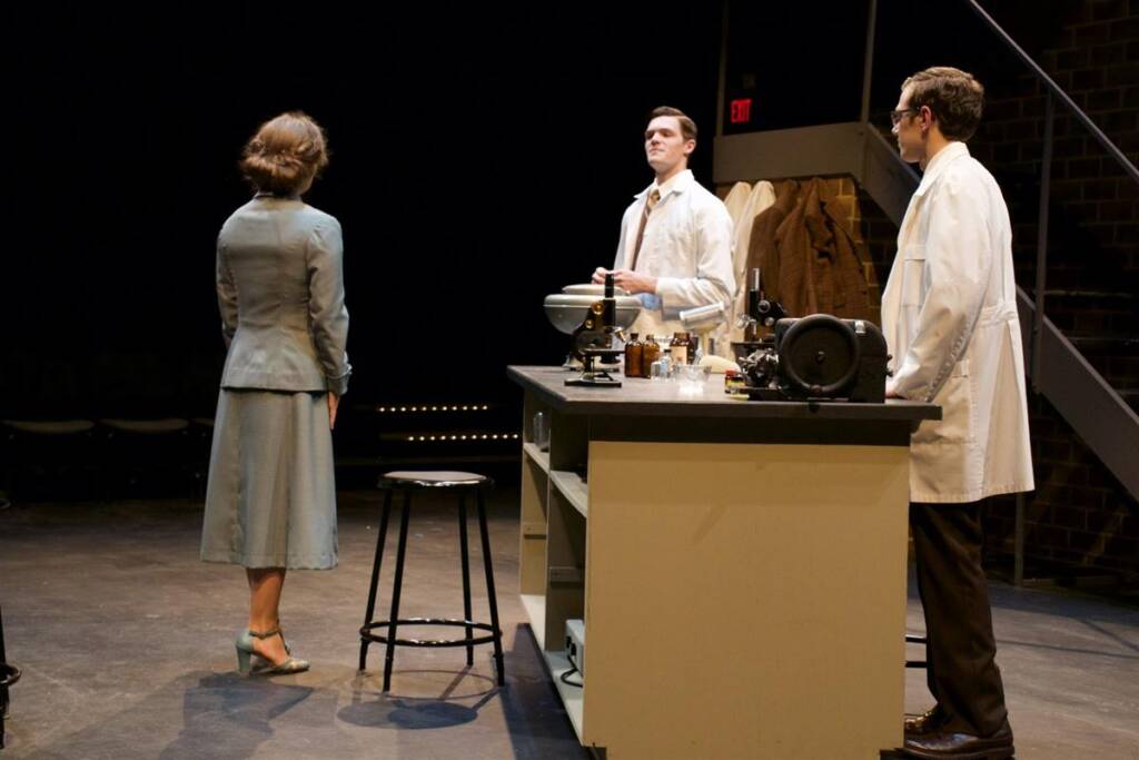 A woman in a teal suit faces two men in white lab coats at a laboratory table. One man holds a bowl, and various lab equipment is visible on the table. Coats hang in the background, and a few steps lead up to a darkened area.