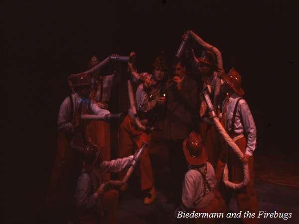 A theatrical scene from "Biedermann and the Firebugs" shows actors wearing orange pants and red helmets, holding hose puppets. The stage is dimly lit, creating a dramatic and intense atmosphere.
