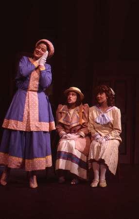 Three actors are on stage in period costumes. One stands to the left in a blue and pink dress, appearing animated. Two sit to the right in complementary outfits, including hats and stripes, watching. The lighting is warm and theatrical.