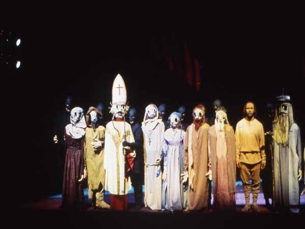 A group of people on stage wearing elaborate masks and costumes. Some are dressed in robes, with one in a white robe and a bishop's hat. The lighting is dim, creating a dramatic atmosphere.