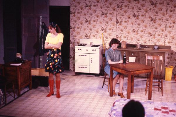 Two people are in a vintage kitchen. One stands with arms crossed, wearing a yellow top and a floral skirt. The other sits at a table with a bottle, wearing a blue dress. A stove and sink are in the background, with floral wallpaper.