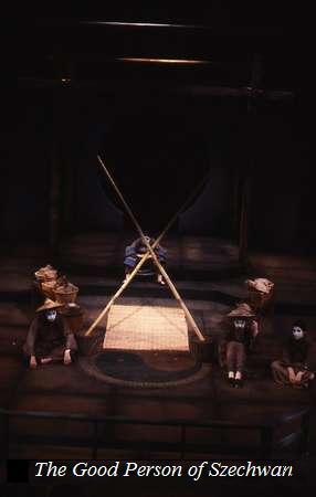 A dimly lit stage of "The Good Person of Szechwan" with actors in dark costumes seated and standing around a triangular bamboo structure. The backdrop is minimal, emphasizing the dramatic scene. Text at the bottom reads "The Good Person of Szechwan.