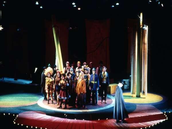 A theater stage with a group of performers in colorful costumes standing in a semi-circle under bright lights. The background features tall wooden pillars. Another person in a cloak stands slightly apart, facing the group.
