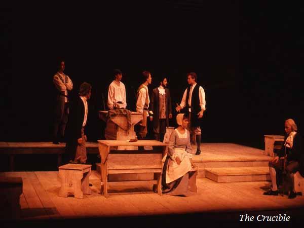 A theater stage with actors in historical costumes performing a scene from "The Crucible." The set includes wooden benches and tables, with dim lighting creating a dramatic atmosphere. The title "The Crucible" is visible in the bottom right corner.