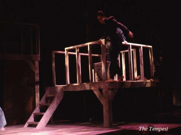A person dressed in dark clothing is climbing over a wooden railing on a dimly lit stage set. The backdrop is shadowy, conveying a theatrical atmosphere. Text at the bottom reads "The Tempest.