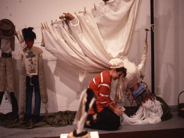 A person in a red and white striped shirt interacts with a seated figure covered in cloth, resembling a laundry scene. White sheets are hung on a line in the background, with another figure in a hat to the left.