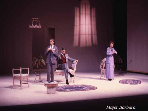 Three actors perform a scene from "Major Barbara" on stage. Two men in suits stand near a small table and white chairs, while a woman in a long blue dress stands to the right. The backdrop features a window casting light.