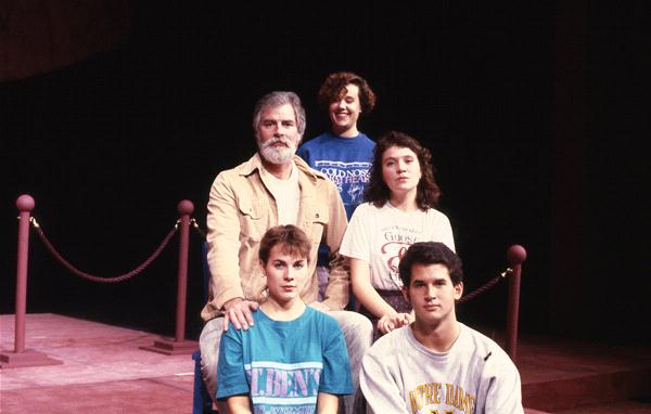 Five people are sitting in two rows on a stage. The older man in the back row, wearing a beige jacket, sits next to a smiling woman in a blue shirt. Three young adults sit in front, all wearing casual shirts. The background is dark with stage ropes visible.