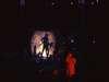 Silhouetted figures pose dramatically in front of a bright, circular light source in a dark setting. A person in a red outfit stands in the foreground, adding contrast to the scene.