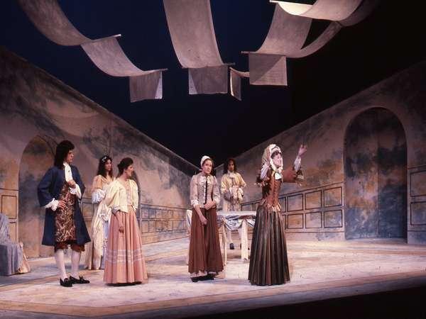 A theatrical scene with six actors in historical costumes on an indoor stage. The set includes curved walls and hanging fabric. The lighting is soft, creating a dramatic atmosphere. The actors engage in what appears to be a lively dialogue or performance.
