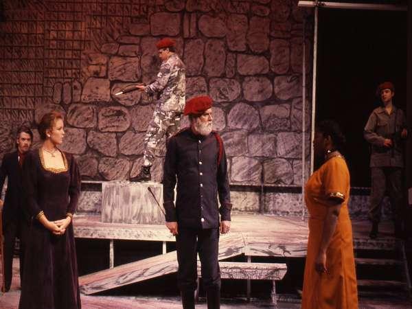 A theatrical stage scene with actors in historical and military costumes. A man with a beard in a black uniform faces a woman in a mustard dress. Another actor in camouflage holds a gun, while two others stand in the background with stone wall props.