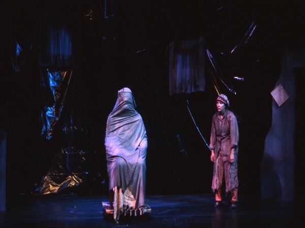 A dimly lit stage features a figure draped in a cloak seated on a stone. Another figure stands nearby, wearing ragged clothes and a hat, gazing at the seated figure. The background is dark with textured elements creating an eerie atmosphere.