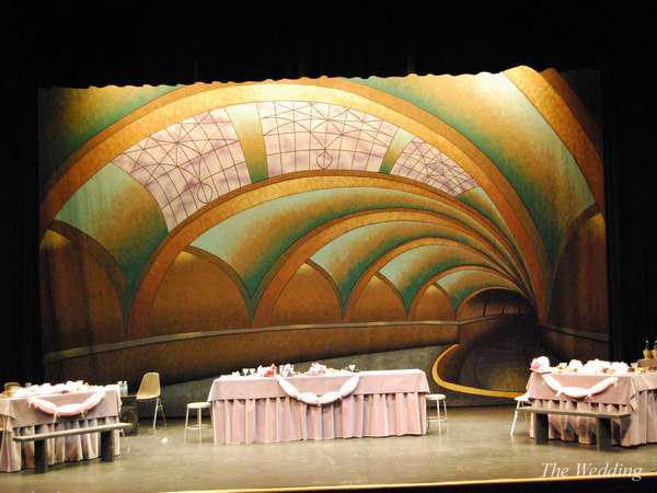 A theater stage set for a wedding scene with a backdrop featuring an abstract, arched design resembling an art deco ceiling. The stage has decorated tables with pink tablecloths and white garlands.