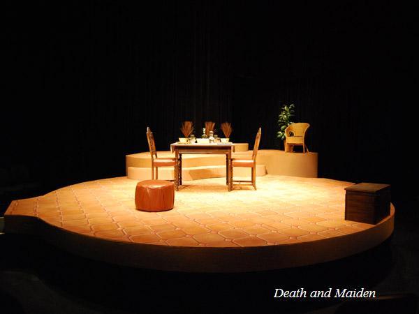 The image shows a theater stage set for "Death and the Maiden." It features a round platform with a wooden table, chairs, a small plant, an ottoman, and a chest. The background is dark, with "Death and Maiden" text in the corner.