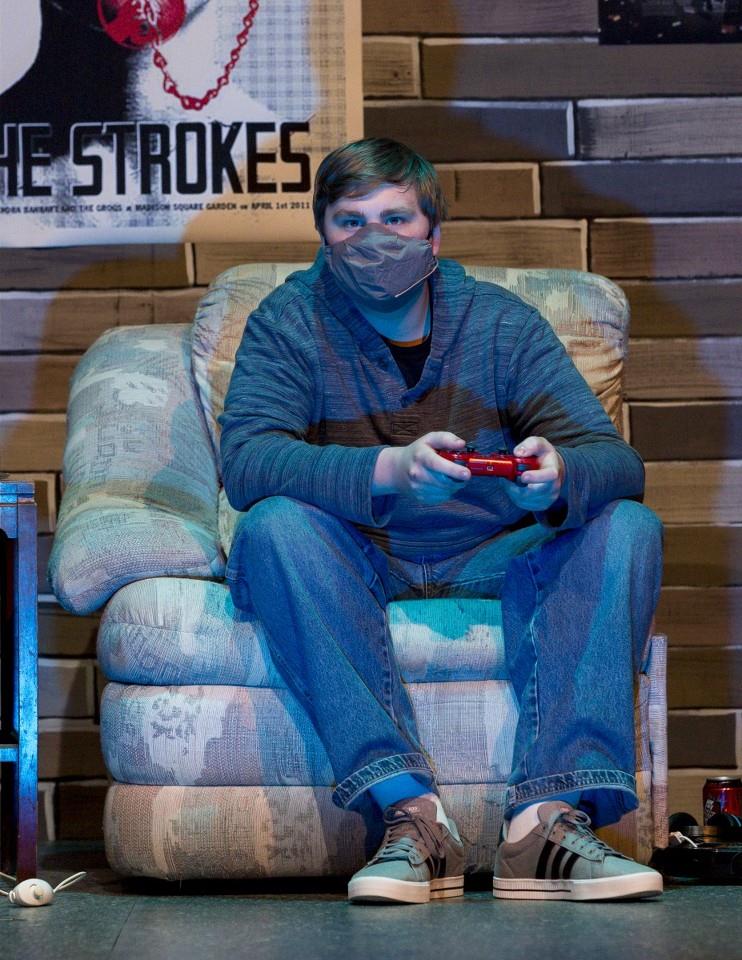 A person seated on a patterned armchair, wearing a gray face mask, blue jeans, and a navy blue hoodie, is holding a red game controller. Behind them is a poster and a wooden wall. Their expression is focused on the game.