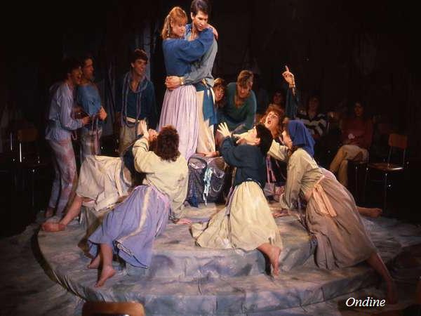 A theatrical performance with actors on stage. A central couple stands embraced on a raised platform, surrounded by others reaching toward them with expressive gestures. The background is dark, and several seated observers are visible. Text: "Ondine.